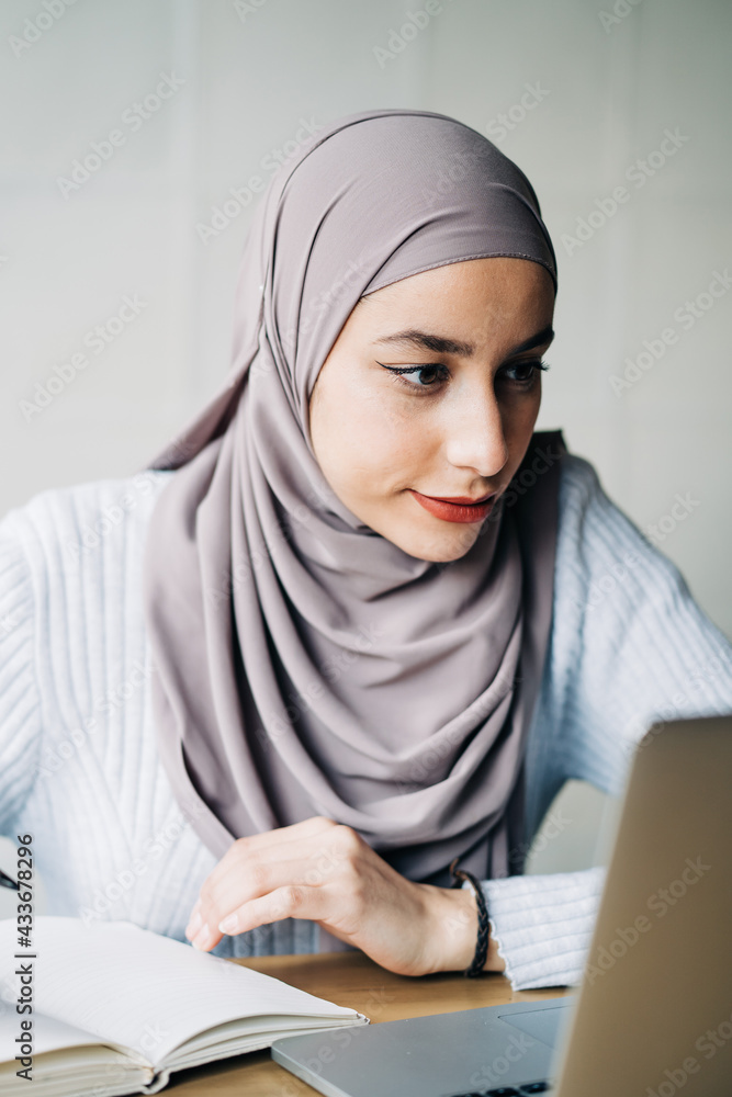Portrait of ethnic female freelancer in hijab using laptop while sitting at table in cafe and working remotely Portrait of ethnic female freelancer in hijab using laptop while sitting at table in cafe and working remotely