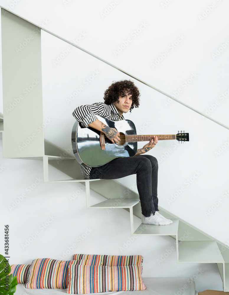 Side view of young hipster male musician playing acoustic guitar while  sitting on staircase and looking at camera in flat Stock-foto | Adobe Stock