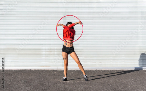 Young tattooed woman in activewear twirling hula hoop while dancing against brick walls with shadows and looking forward in sunlight photo