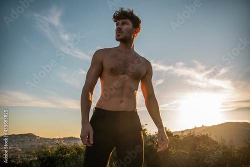 Shirtless male with six pack abs and beard looking away against mountains under cloudy sky in evening