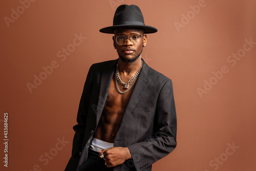 Young masculine unshaven African American male with naked abdomen in jacket looking at camera on brown background photo