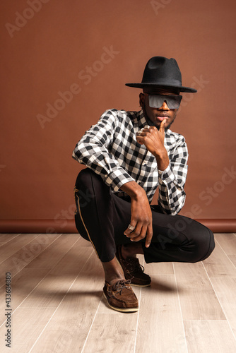 Young cool African American male model in checkered shirt and hat looking at camera while crouched on brown background