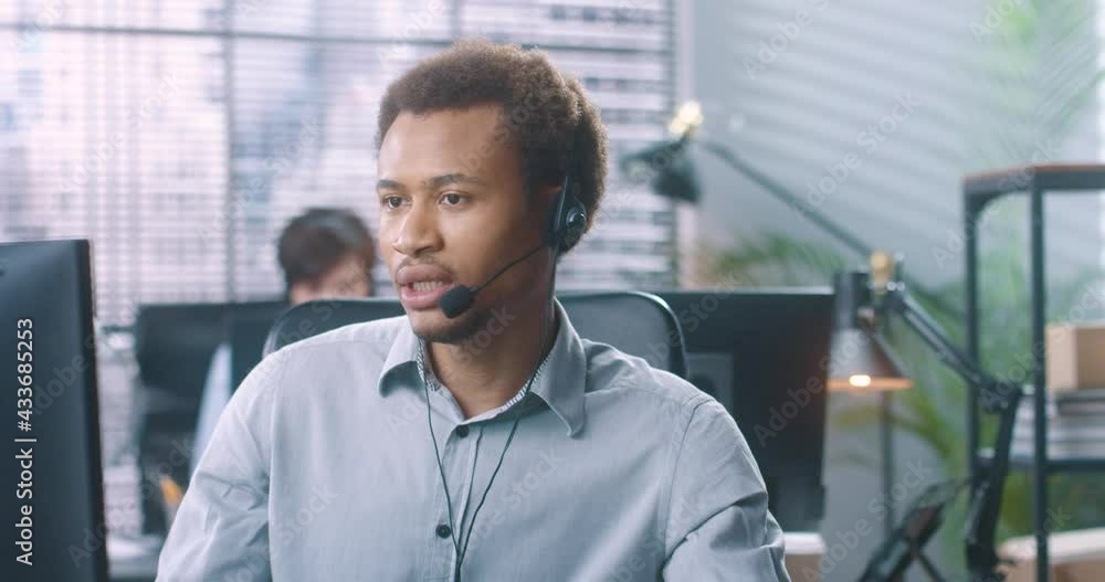 Close up portrait of serious young African American male worker speaking in headset on video call on computer sitting at table at office. Handsome man employee talk with client on online call Close up portrait of serious young African American male worker speaking in headset on video call on computer sitting at table at office. Handsome man employee talk with client on online call