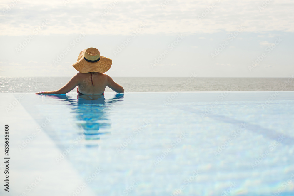 Beautiful woman in hat relaxing at edge in blue pool, enjoying summer vacation and calm sea view. Slim young female relaxing at tropical resort in swimming pool. Travel and Holidays. Space for text - obrazy, fototapety, plakaty 