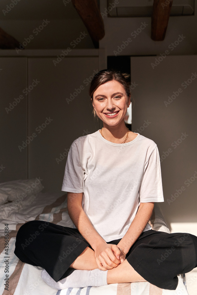 Full body of positive young lady in casual outfit and socks sitting on comfortable bed with crossed legs and smiling Full body of positive young lady in casual outfit and socks sitting on comfortable bed with crossed legs and smiling