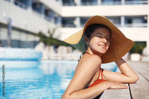 Enjoying summer vacation. Beautiful happy woman in hat relaxing in pool water. Stylish slim young female sunbathing and resting at swimming pool edge at tropical resort. Holidays and travel