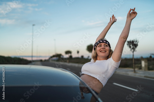 Young female in casual wear and headband with American flag print leaning out of car window and raising hands while enjoying freedom during road trip at sunset photo