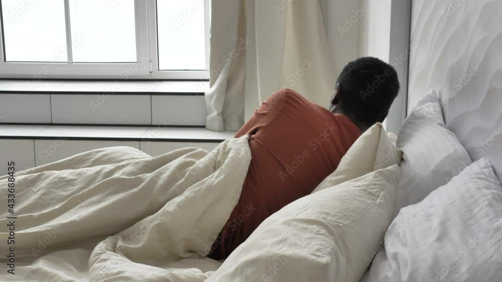 Positive African-American guy in pajamas awakes and stretches in comfortable bed in light bedroom in sunny morning closeup Positive African-American guy in pajamas awakes and stretches in comfortable bed in light bedroom in sunny morning closeup