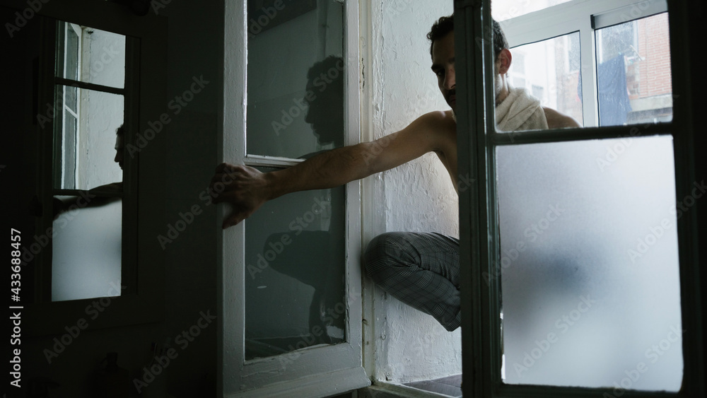 Pensive male sitting on knees on windowsill at home