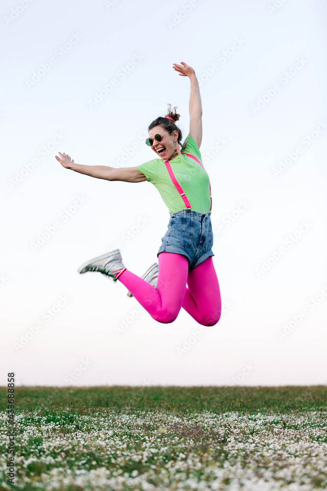 Full body delighted young female wearing shorts and bright pink tights jumping merrily with arm raised on lush grassy glade in nature Full body delighted young female wearing shorts and bright pink tights jumping merrily with arm raised on lush grassy glade in nature