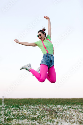 Full body delighted young female wearing shorts and bright pink tights jumping merrily with arm raised on lush grassy glade in nature