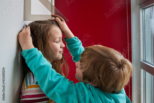 Brother helping sister with measuring her height with ruler and pencil near wall photo