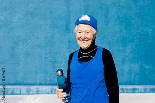 Cheerful mature sportswoman in activewear and cap standing with water bottle in hand against blue wall in sunny outdoor training center and looking away photo