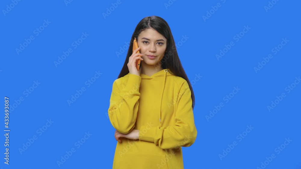Portrait of pretty brunette talking on a smartphone. Young woman with long hair in yellow sweatshirt posing in studio with blue screen background. Close up. Slow motion ready 59.94fps. Portrait of pretty brunette talking on a smartphone. Young woman with long hair in yellow sweatshirt posing in studio with blue screen background. Close up. Slow motion ready 59.94fps.