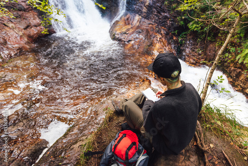 unrecognizable person sitting on a rock in front of a waterfall in the forest taking notes in his notebook during a break on his mountain path. hiking woman. activities in the nature.