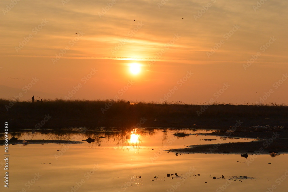 S+unset view from Mavisehir, Karsiyaka, Izmir. Reflection of the sun with beautiful orange and yellow colors and reeds. The people in silhouette are enjoying scenery and nature. S+unset view from Mavisehir, Karsiyaka, Izmir. Reflection of the sun with beautiful orange and yellow colors and reeds. The people in silhouette are enjoying scenery and nature.