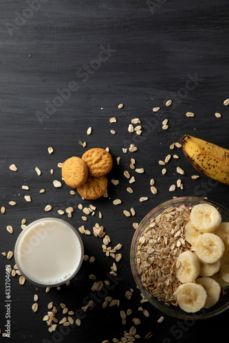Desayuno saludable de avena con plátano, un vaso de leche y unas galletas sobre la mesa de color negro.