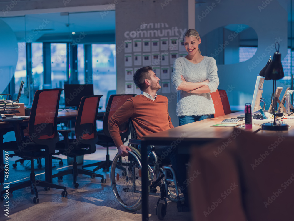 Obraz premium Businessman in a wheelchair in modern coworking office space working late night in office. Colleagues in background. Disability and handicap concept. 