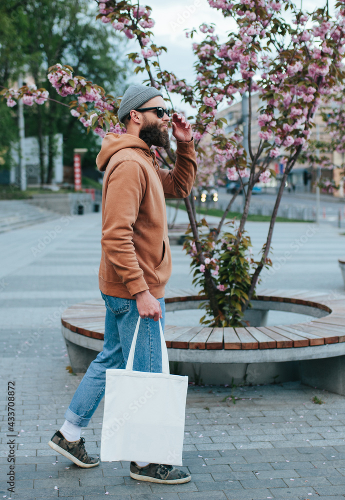 Street style portrait of young hipster man wearing brown blank hoodie with  white blank textile eco shopping bag or tote bag. Mock up for design Stock  Photo