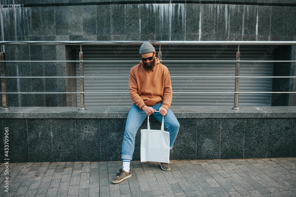 Street style portrait of young hipster man wearing brown blank hoodie with  white blank textile eco shopping bag or tote bag. Mock up for design Stock  Photo