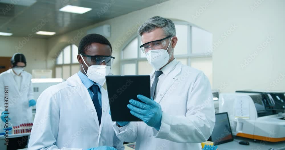 Close up portrait of Caucasian senior man doctor talking to African American young male medical scientist in mask standing in clinic lab and typing on tablet discussing treatment results, medicine Close up portrait of Caucasian senior man doctor talking to African American young male medical scientist in mask standing in clinic lab and typing on tablet discussing treatment results, medicine