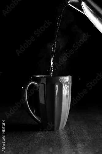 A mug with hot steaming water being poured from a metal kettel in front of a black background on a dark wooden surface. photo