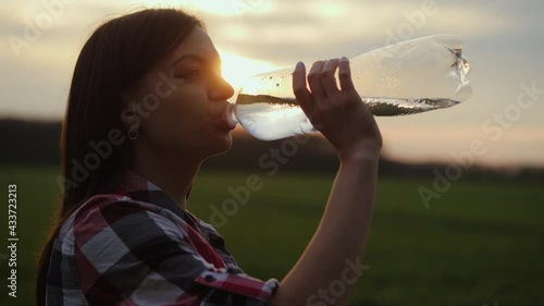 Woman quenches her thirst in a field at sunset. Silhouette of a girl drinks water from a plastic bottle, sun rays through the bottle