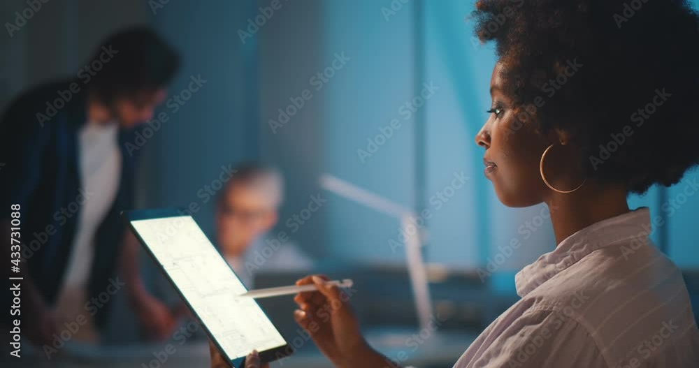 Professional afro-american architect studying blueprint on digital tablet in office working late