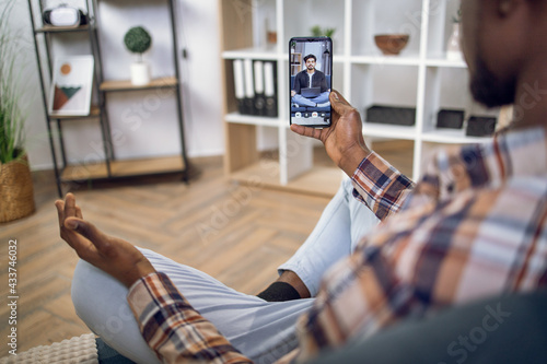 View from shoulder of afro american man holding smartphone and meditating on couch during video call with muslim psychologist. Online consultation during pandemic.