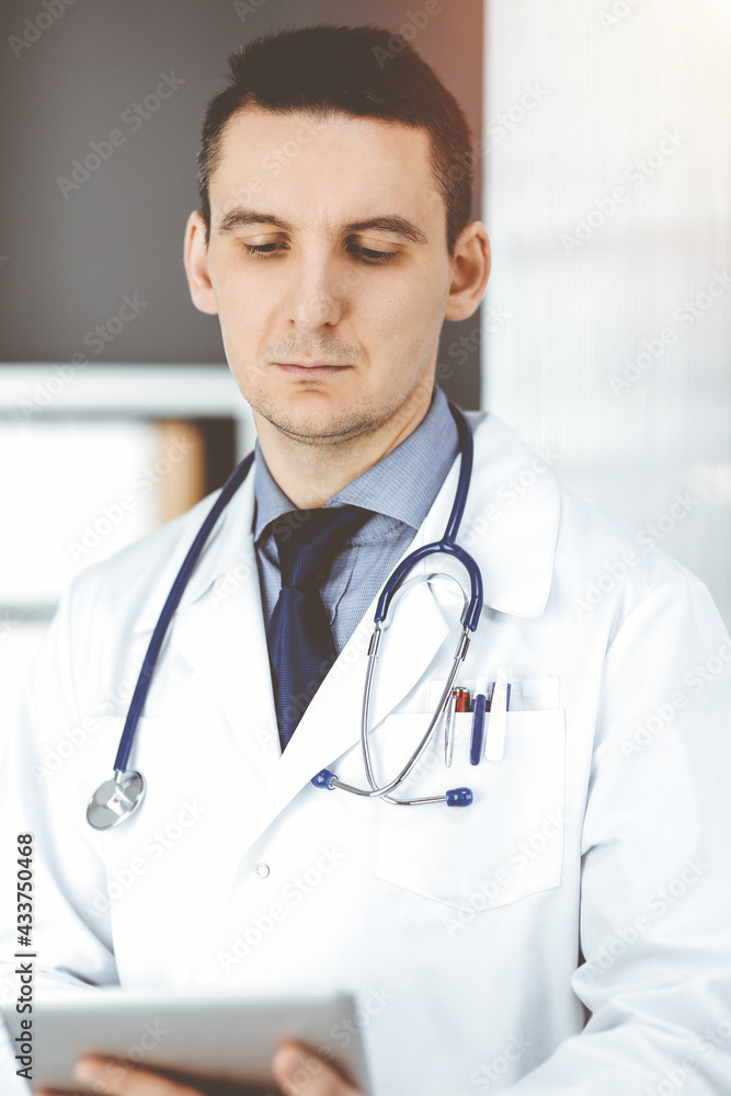 Friendly male doctor using tablet computer in clinic near his working place. Perfect medical service in hospital. Medicine and healthcare concept Friendly male doctor using tablet computer in clinic near his working place. Perfect medical service in hospital. Medicine and healthcare concept
