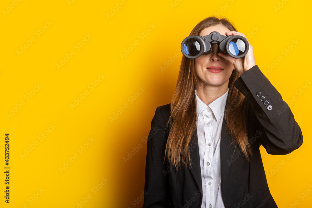 young business woman looking through binoculars on a yellow background - obrazy, fototapety, plakaty 