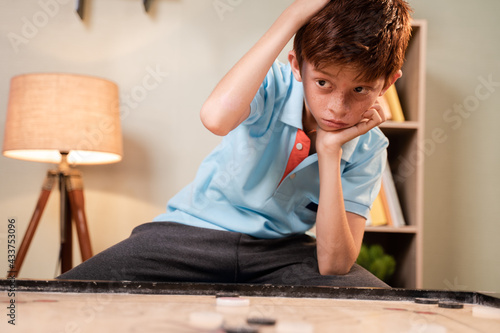 Overthinking Confused Caucasian kid scratching head to decide which coin to hit while playing carrom board - concept of mental illness and development