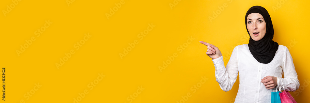 Surprised Young Muslim woman in hijab holds shopping bags and points finger to the side against yellow background. Banner Surprised Young Muslim woman in hijab holds shopping bags and points finger to the side against yellow background. Banner