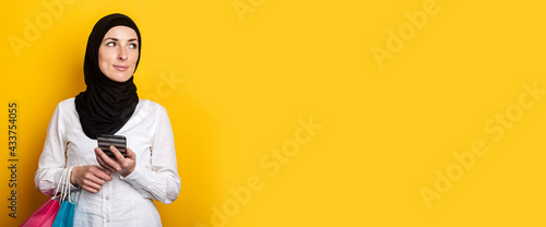 Cute Young Muslim woman in hijab holds phone and shopping bags on yellow background. Banner © Alex