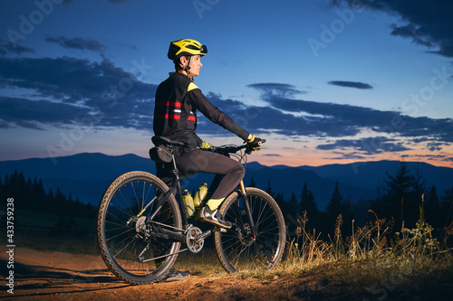 Side view of young man on bicycle under blue night sky with clouds. Male bicyclist in safety helmet riding bicycle on the mountain trail in the evening. Concept of sport, biking and active leisure.
