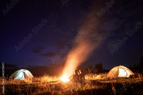 Horizontal snapshot of couple of tourists spending time together in camping. Young man and woman, sitting close near campfire in the evening outdoors in nature. Glowing tents on the sides