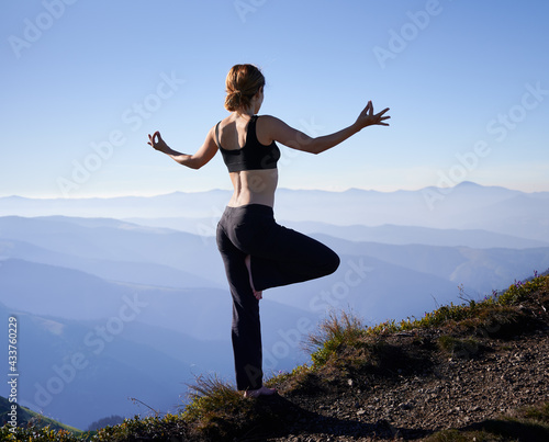 Back view of sporty woman doing yoga exercise and standing on one leg in mountains. Concept of harmony with nature.