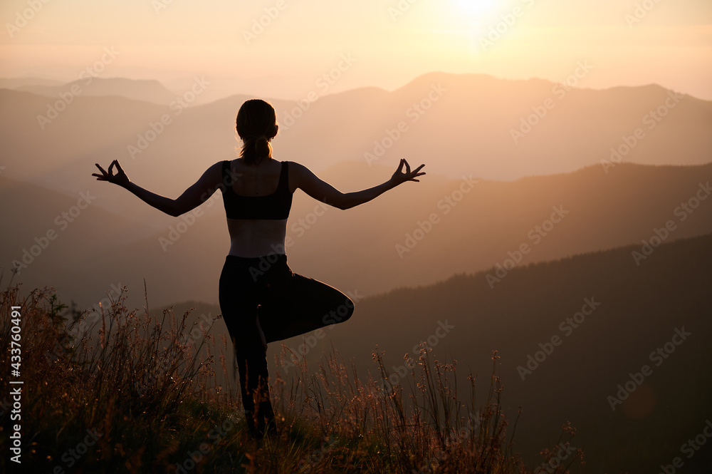 Back view of woman practicing yoga on background of evening mountains. Meditating female is balancing on one leg after sunset. Concept of exercise.