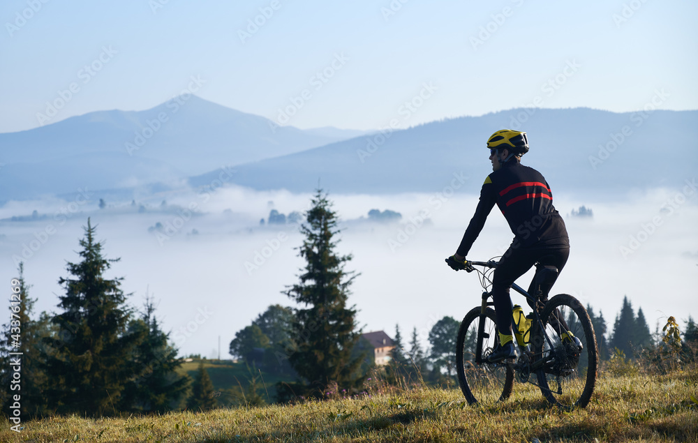 Silhouette of man cyclist in cycling suit riding bike on grassy hill. Male bicyclist enjoying the view of majestic mountains during bicycle ride. Concept of sport, bicycling and nature. Silhouette of man cyclist in cycling suit riding bike on grassy hill. Male bicyclist enjoying the view of majestic mountains during bicycle ride. Concept of sport, bicycling and nature.