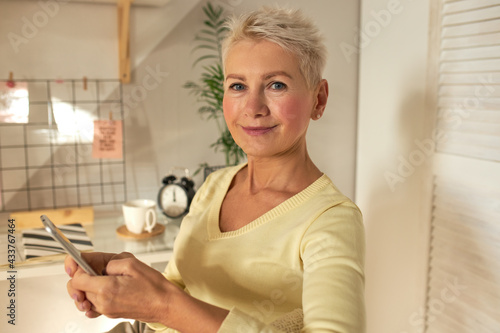 Indoor image of beautiful mature Caucasian businesswoman with short hairstyle using generic cell phone for distant work checking email, typing text message, looking at camera with confident smile