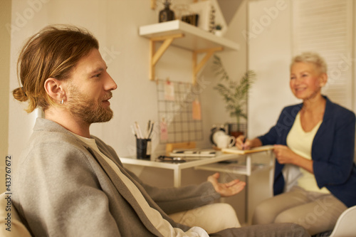 Portrait of handsome young man with stylish hairdo and beard having conversation with his female psychologist. Blonde woman coach writing down information during session with stylish male