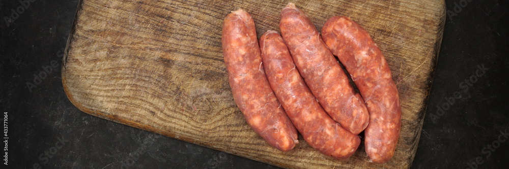 Raw Sausages In Natural Casing. Stuffed Sausages on Old Wooden Cutting Board and Fork, Overhead View. Uncooked Sausages For Grilling or Frying on Wood Board And Black Table in Background, Top View. Raw Sausages In Natural Casing. Stuffed Sausages on Old Wooden Cutting Board and Fork, Overhead View. Uncooked Sausages For Grilling or Frying on Wood Board And Black Table in Background, Top View.