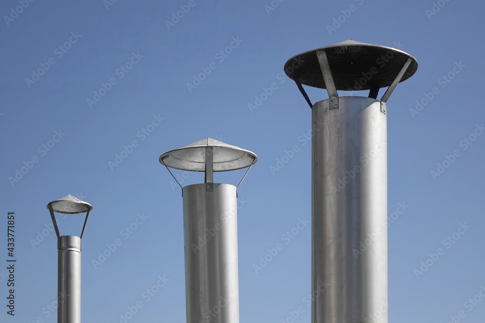 Three Modern New And Clean Galvanized Chimney Pipe Or Zinced Stovepipe On House Roof Close Up View Against The Blue Sky Background Isolated. Three Modern New And Clean Galvanized Chimney Pipe Or Zinced Stovepipe On House Roof Close Up View Against The Blue Sky Background Isolated.