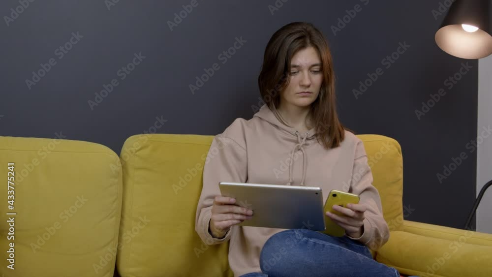 Attractive long-haired brunette girl chatting and messaging sitting on sofa in cozy stylish living room. Young lady holds tablet and smartphone listening to music, and communicating on social networks Attractive long-haired brunette girl chatting and messaging sitting on sofa in cozy stylish living room. Young lady holds tablet and smartphone listening to music, and communicating on social networks