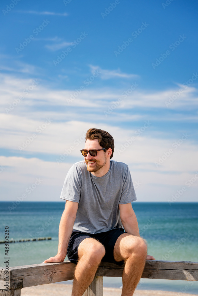 Smiling young man in sunglasses, resting on a wooden railing by the beach. Smiling young man in sunglasses, resting on a wooden railing by the beach.