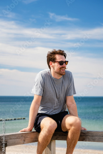Smiling young man in sunglasses, resting on a wooden railing by the beach. © ryszard filipowicz