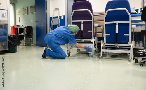 Woman from the cleaning service of a hospital crouching cleaning the wheels of a patient chair in the operating room beforehand. Sanitary concept