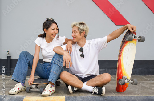 Asian couple resting after skateboarding. A young man in a white T-shirt held his hand on the surfskates board while talking to his girlfriend. The woman smile happily with sunglasses on her head. photo