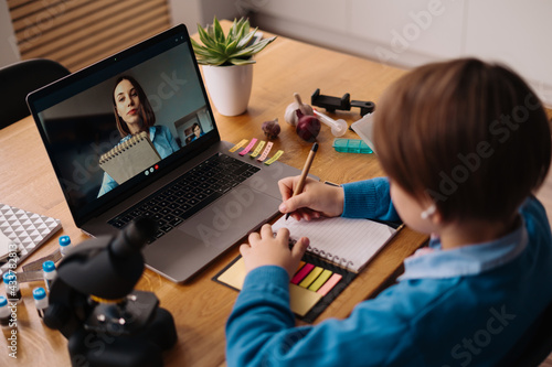 A Preteen boy uses a laptop to make a video call with his teacher. The Screen shows an online lecture with a teacher explaining the subject from class.