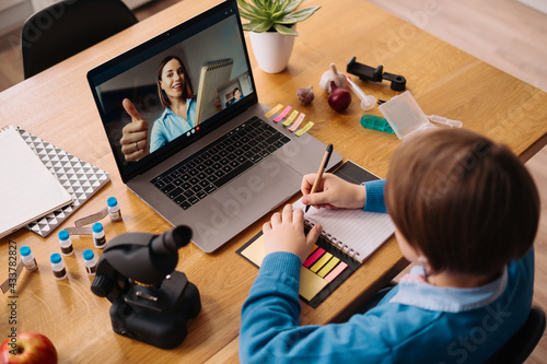 A Preteen boy uses a laptop to make a video call with his teacher. The Screen shows an online lecture with a teacher explaining the subject from class.